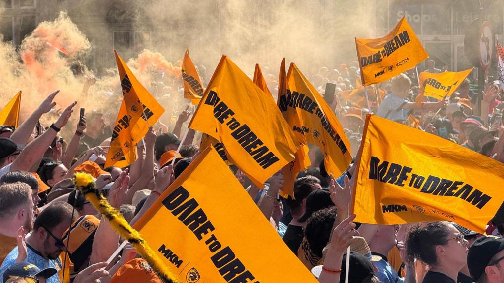 Hull City fans waving amber "dare to dream" flags in a city square with amber flares going off