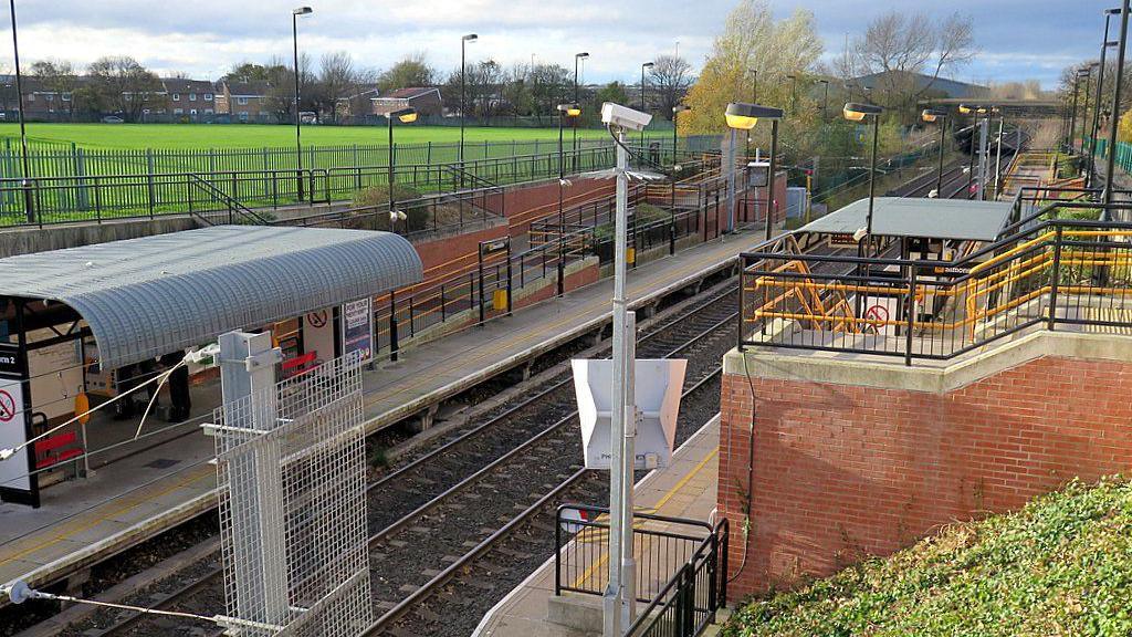 A picture of the Metro station. It is outdoors and has a curved metal roof on one platform. The opposite platform has a flat roof and there are stairs leading down to it.