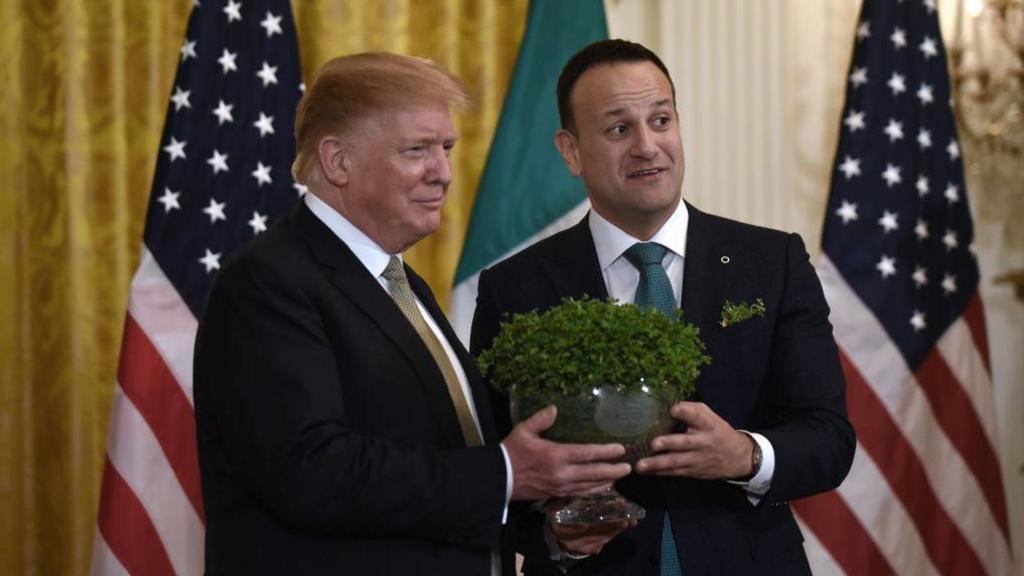 A man with blonde hair in a black suit with shirt shirt and gold tie being handed a glass bowl of green shamrocks by a man with short brown hair in a navy suit white shirt and green tie. They are stood in front of several USA flags - white and red stripes with blue and white stars.