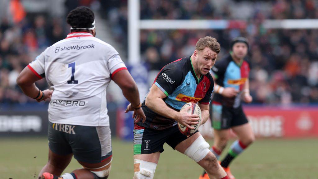 Rugby player in multi-coloured strip side-stepping with the ball in hand as an opponent in white and grey wearing the number 1 watches him and prepares for the tackle