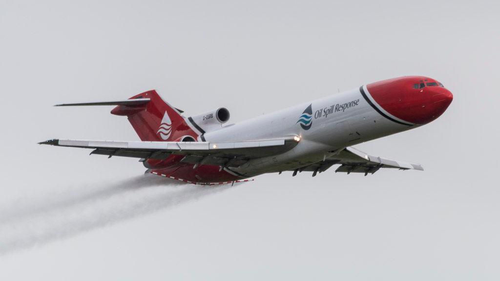 A Boeing 727 tri-jet flying on a grey day. It is painted with red and white livery. It is spraying a liquid from red and white booms attached to the back of the aircraft.