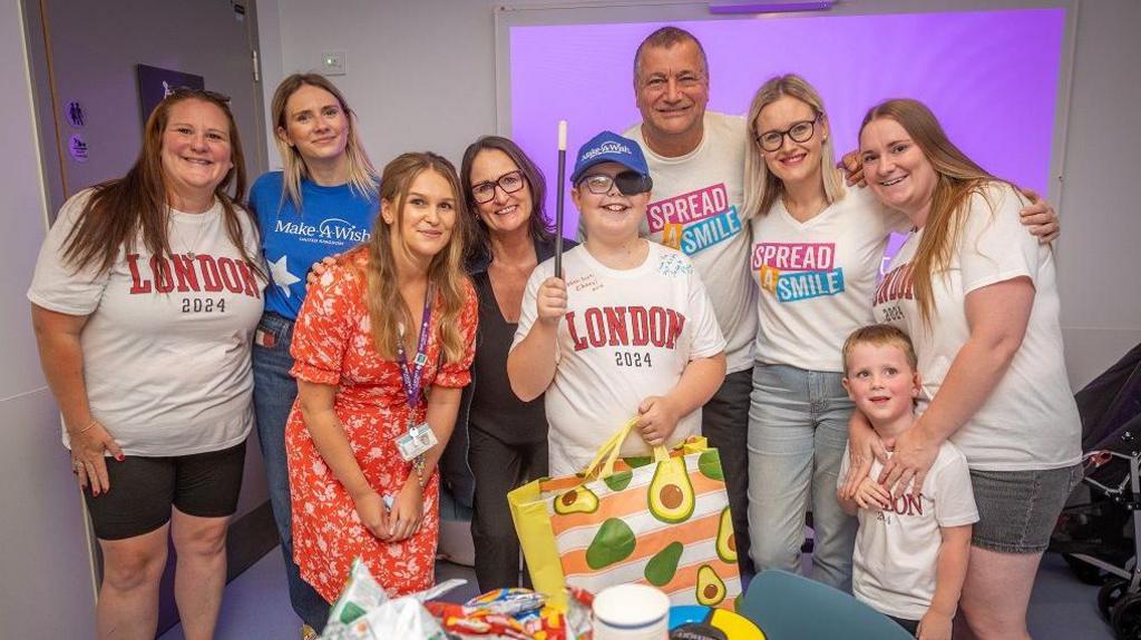 A group of people smiling with their arms round each other. Some of them are wearing t-shirts saying 'London 2024' on them, while others' say 'Spread a smile'. At the front stands Jacob, a 10-year-old boy with glasses and holding a magic wand. He's wearing a cap saying 'Make-A-Wish' and smiling.