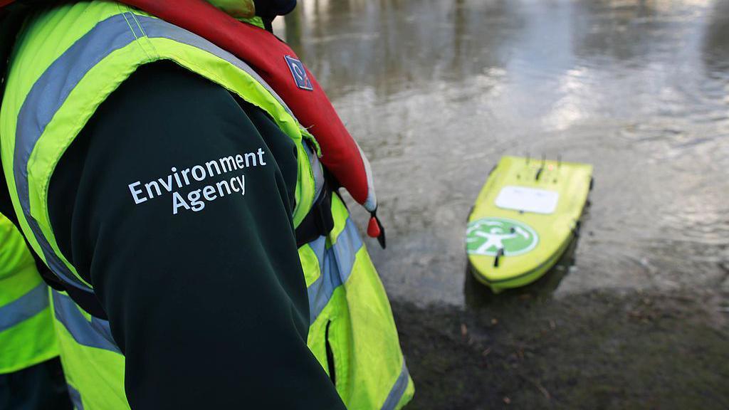 An unidentifiable man standing next to a river wearing a life jacket, a high-visibility vest and a green fleece saying "Environment Agency" on the shoulder. There's a green kayak in the water with the Environment Agency logo on it.