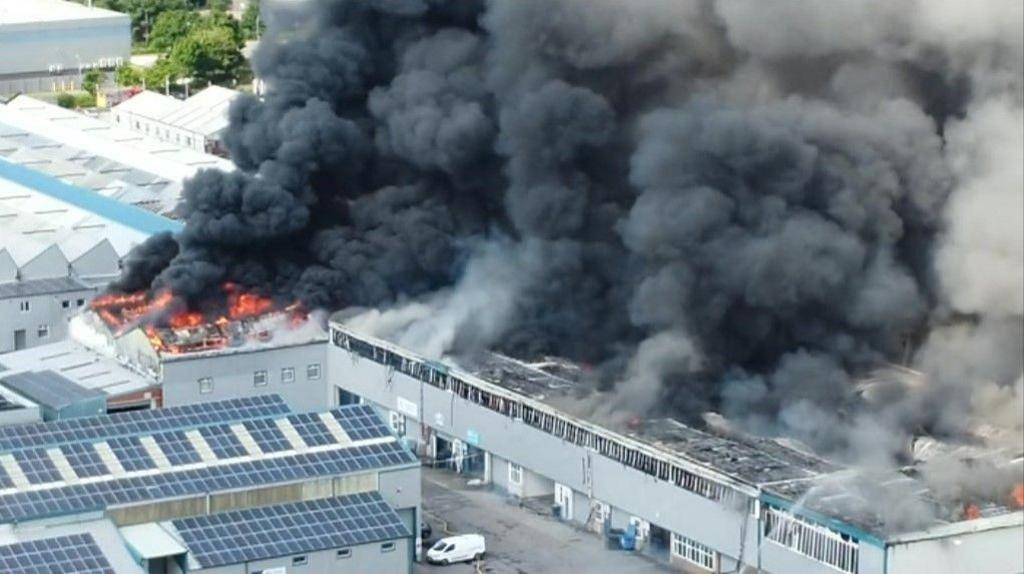 Plumes of dark grey smoke rise over an industrial park, with flames visible on a building's roof