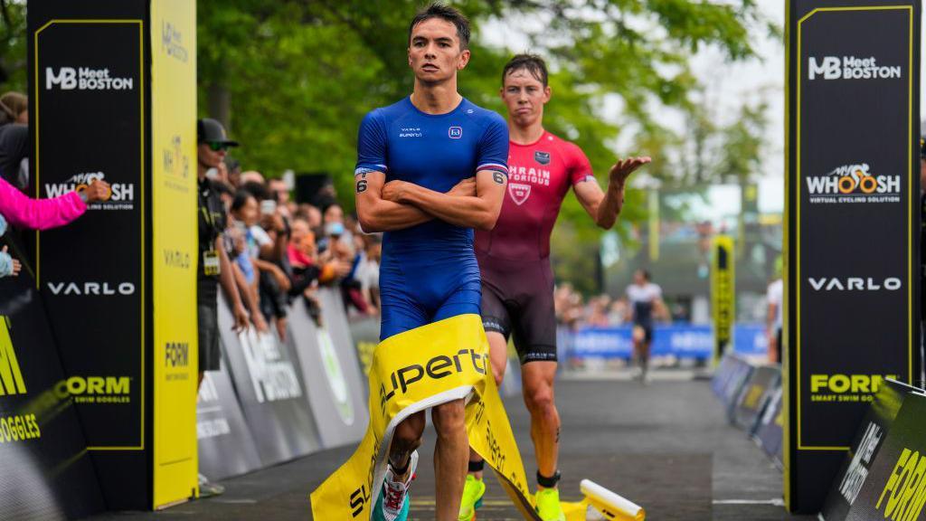 Alex Yee crosses his arms in a 'cold' celebration as he win a Boston Triathlon, with Hayden Wilde gesticulating behind him