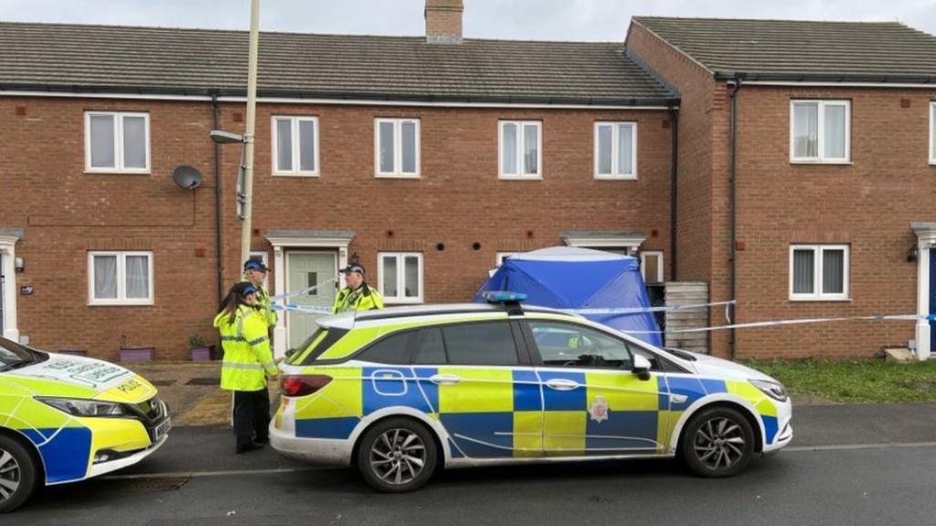 Two police cars parked outside of a series of terraced new build houses. Three police officers are on the scene, where a police cordon and a blue tent have been erected.