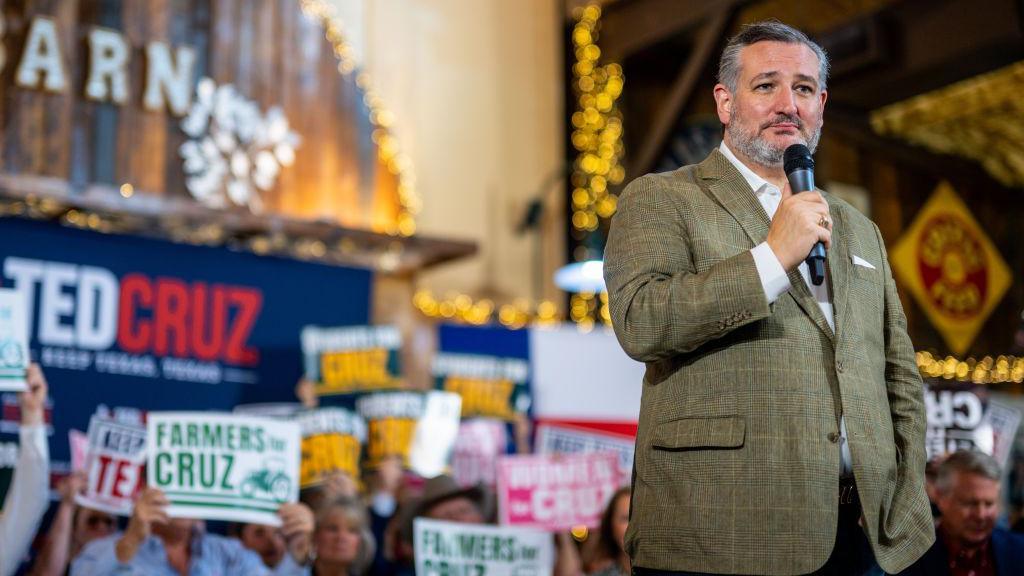 Ted Cruz, wearing a grey tweed blazer over a white dress shirt, and holding a microphone near his mouth, pauses during a speech to supporters holding 'Farmers for Cruz' signs