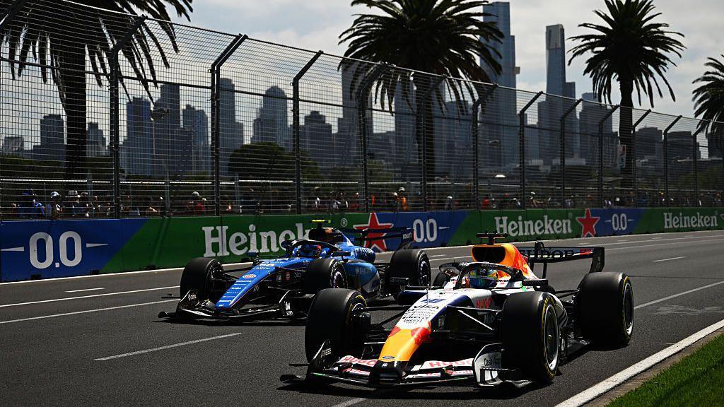 Racing Bulls' Arvid Lindblad slightly ahead of Williams' Carlos Sainz during first practice for the Australian Grand Prix. Stewards watching on, high-rise buildings and palm trees can be seen in the background