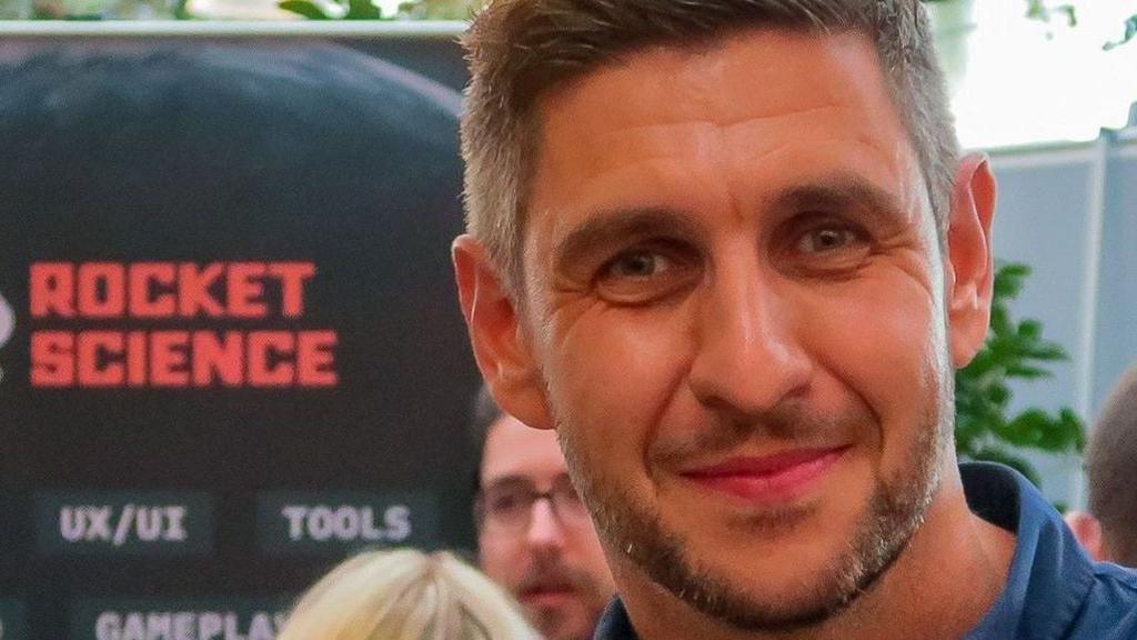 Tom Daniel smiles into the camera at an event, in front of his company's logo. He is wearing a blue shirt and has short, grey hair and a stubbly beard.