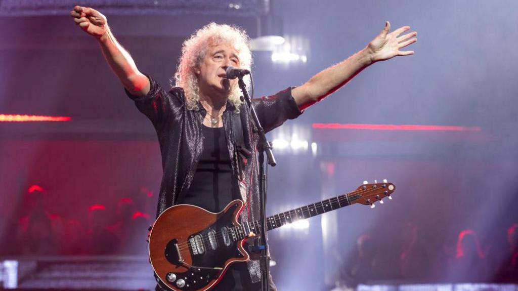 Brian May stands with arms outstretched during a concert, wearing his guitar and a black outfit with audience and stage lightning seen in background