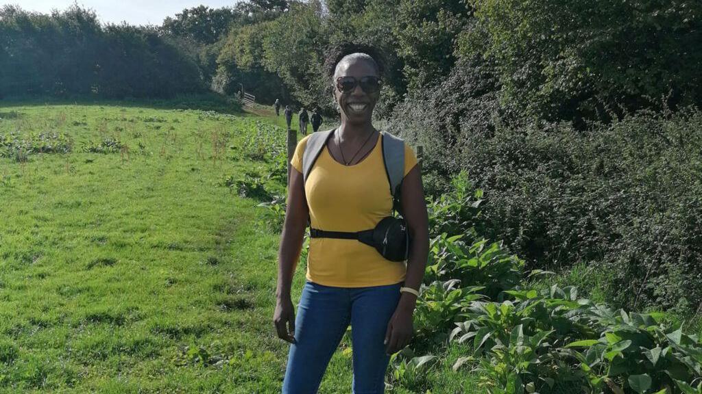 Sophia Brown smiling on a sunny day, standing on a walking trail which cuts through a field. She is wearing blue skinny jeans, a yellow t-shirt, and large sunglasses. 