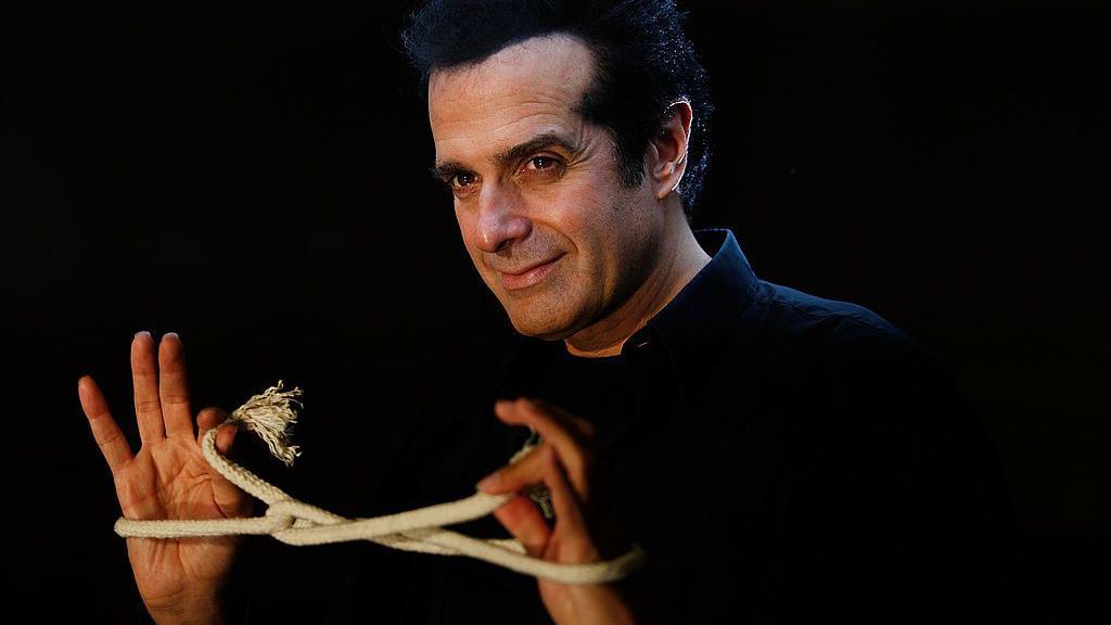 Magician David Copperfield stands in front of a black background. He is wearing a black shirt, and is looking off to the side of the camera, smiling. He has a rope tied in an intricate knot around both hands, and is holding the ends.