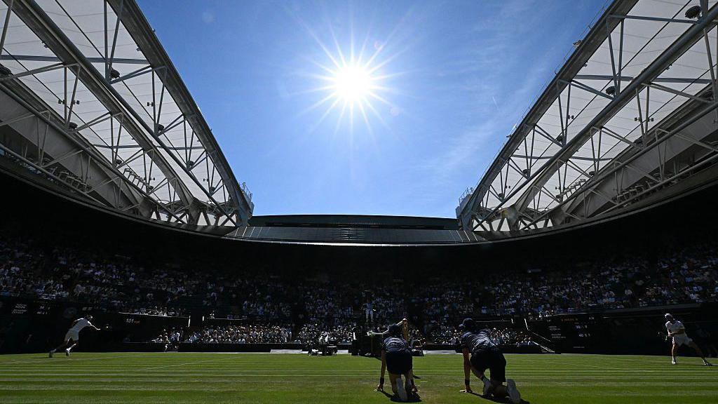 The court can be seen with players in action and with the roof open. There is a bright blue sky and bright sun
