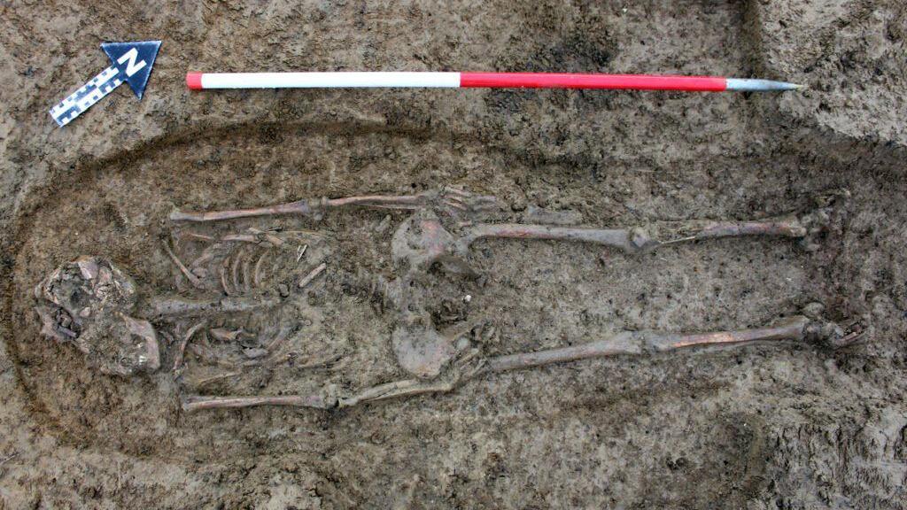 The skeleton of a Roman woman lies in the dirt. There is archaeological equipment beside her, including a red and white measuring pole beside her and a blue arrow with an N on it, pointing away from her.