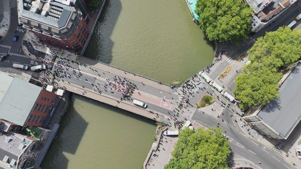 A drone shot above a bridge over a river in Bristol with people scattered on both sides. While all people look tiny, a number of police in high-viz can be spotted.