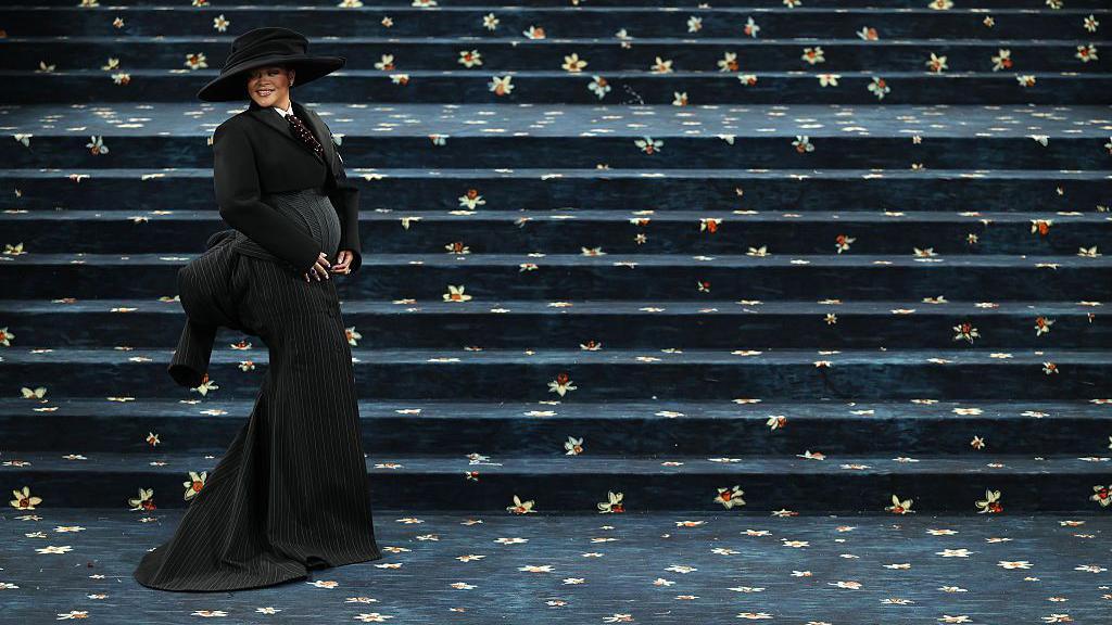 Rihanna smiles and cradles her baby bump as she appears on the red carpet, which is actually navy with daffodils, for the 2025 met Gala. She is wearing a wide-brimmed hat and a floor-length tailored outfit.