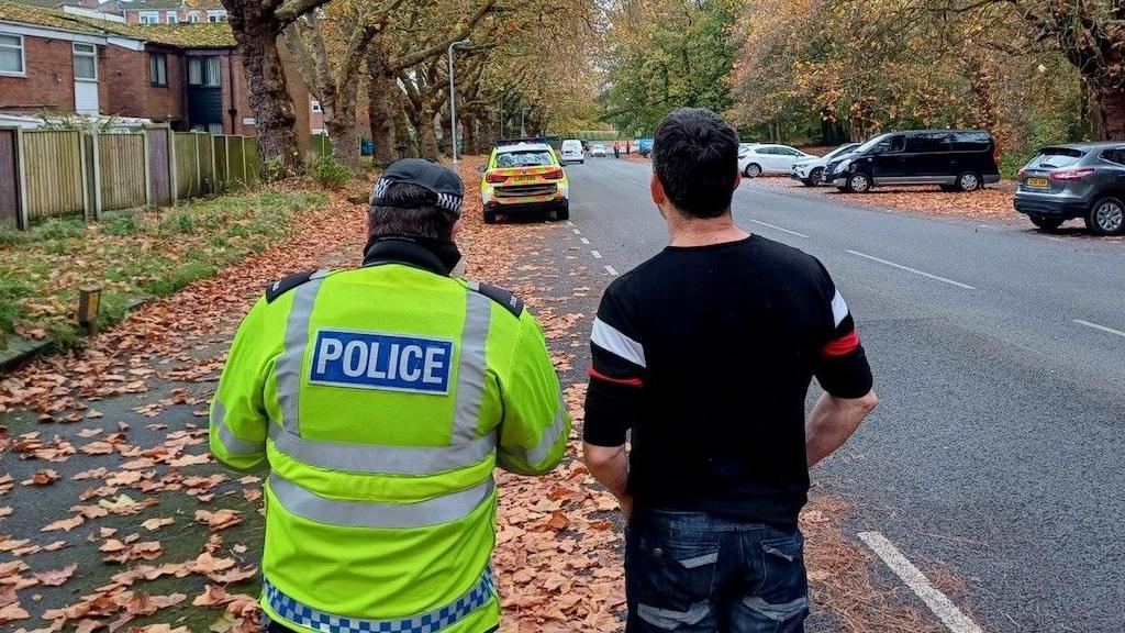 A police officer in a reflective yellow jacket stands next to a man in a black t-shirt. They are facing away from the camera towards the back of a police car parked further down a road, which is littered with fallen leaves