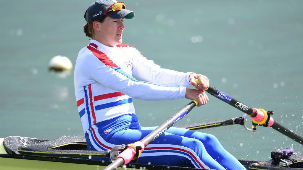 Katherine Grainger sits in a rowing boat as she trains in Redgrave Pincent Rowing Lake. The boat is a pale green, with a large black seat on it. Dame Grainger sits side on to the camera, she wears a dark blue cap, with green under the peak of the cap and large orange mirrored sports sunglasses. Her light brown hair is tied back in a ponytail and she wears a Team GB training suit, consisiting of a whit elong sleeved top, with red and blue stripe detailing down the arms, around her waist and down the sides of her torse - and blue leggings, with white, red and blue striped detailing down the side of her leg. She is holding onto two oars and she rows through the water.