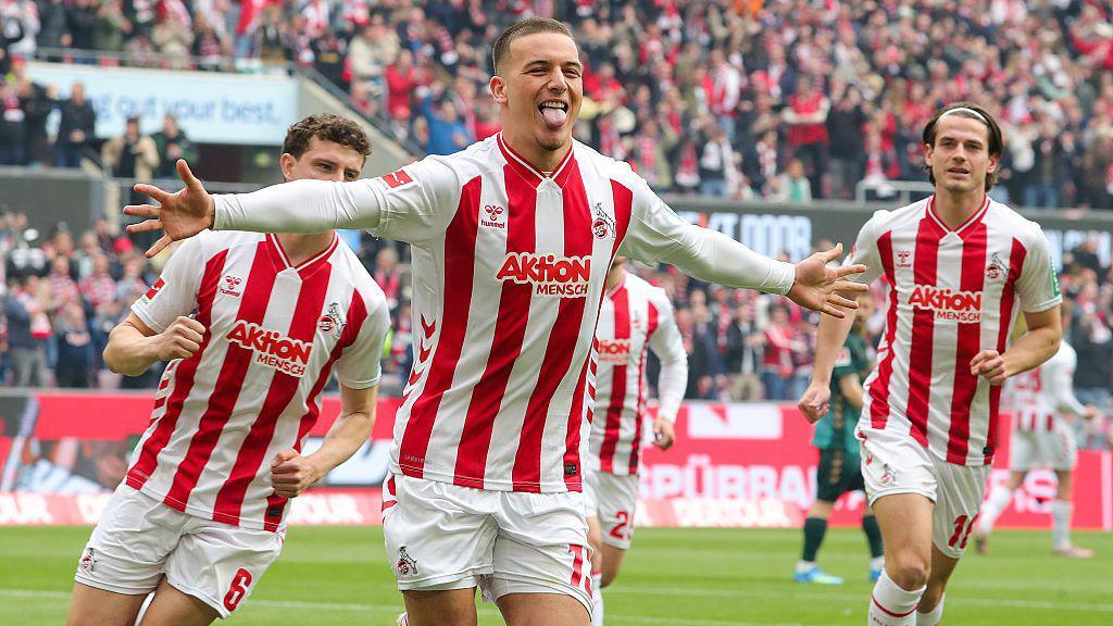 Köln players celebrating a goal against Werder Bremen in their last match. They are wearing red and white striped shirts with white shorts and socks.