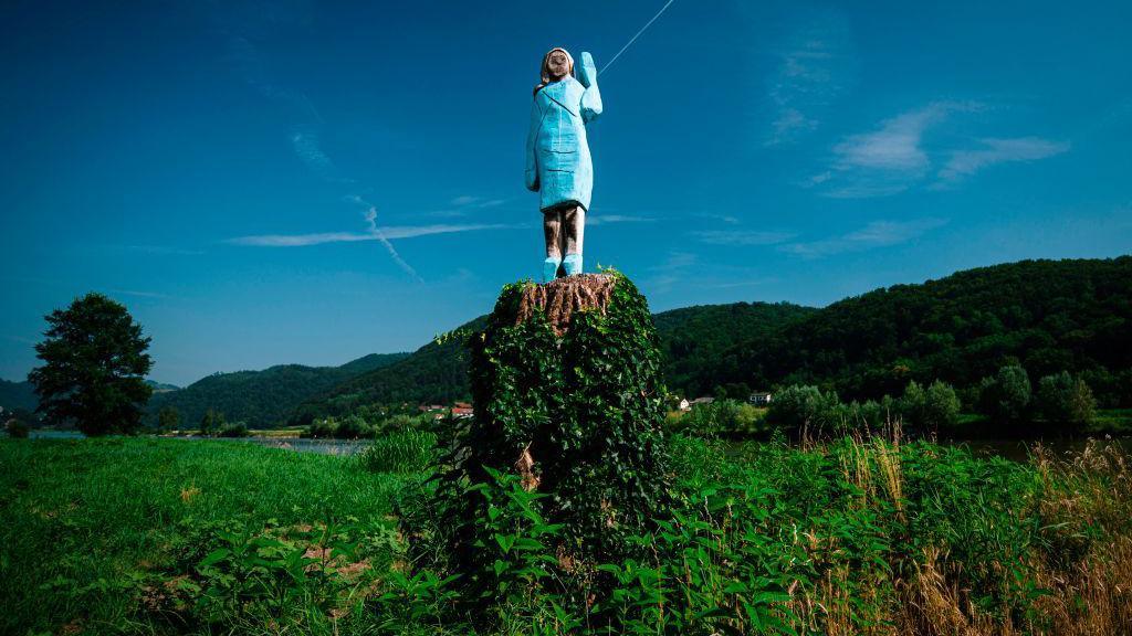 A wooden statue of Melania Trump pictured on top of a tree stump in a green field. This version of the statue has a blue dress and shoes