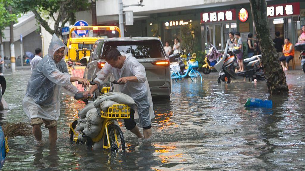 People wearing waterproof ponchos push bicycles loaded with sandbag through shin-deep floodwaters as cars and trucks drive through in the background 