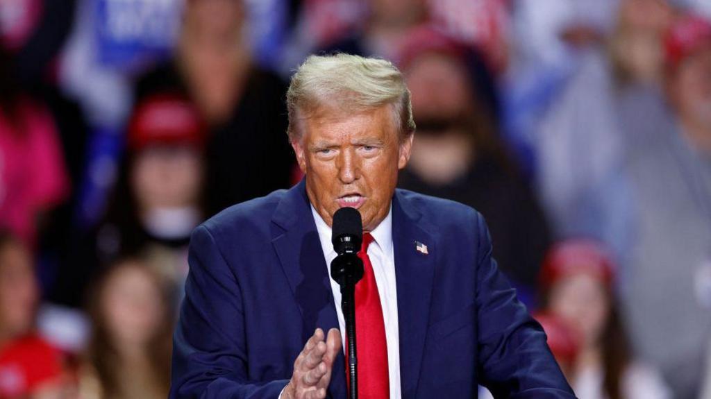 Donald Trump wearing a navy jacket, white shirt and red tie, talking into a microphone and gesticulating with his hand 