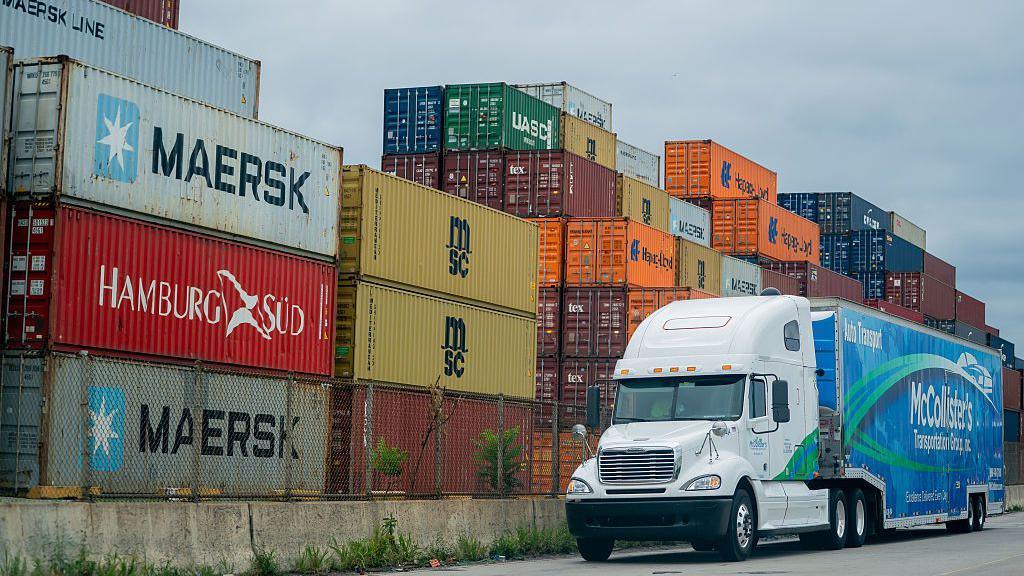 A blue and white McCollister's Transportation Group tractor trailer drives past a pile of shipping containers at the Port of Baltimore in Baltimore, Maryland, US, on Friday, Aug 1, 2025. 