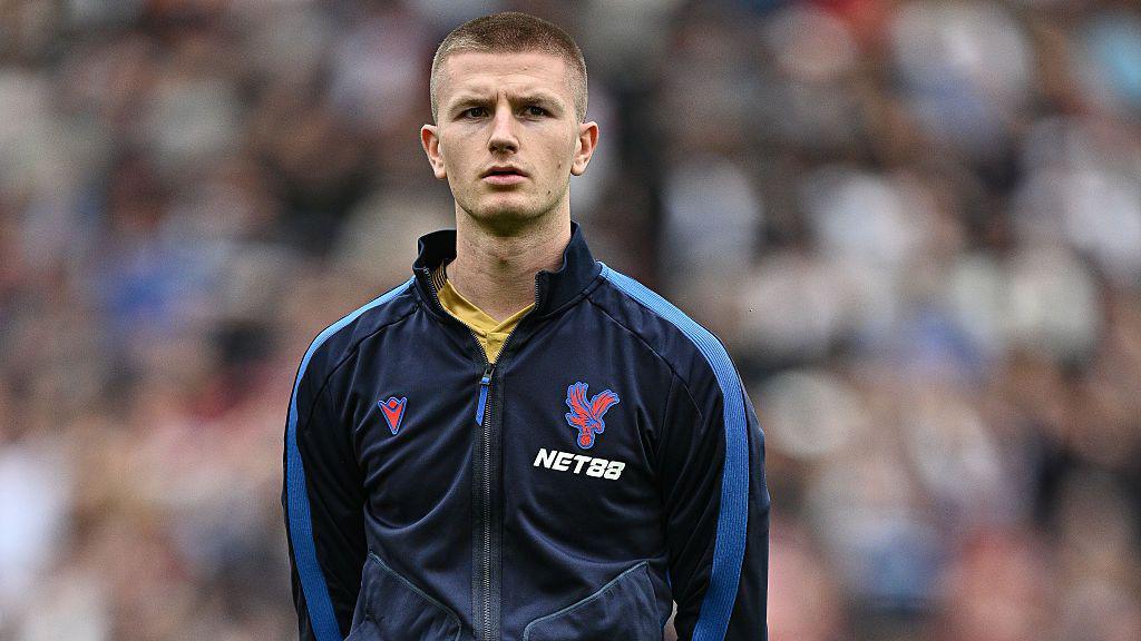 Adam Wharton, wearing a Crystal Palace training top, lining up before a match, with the crowd a blur behind him