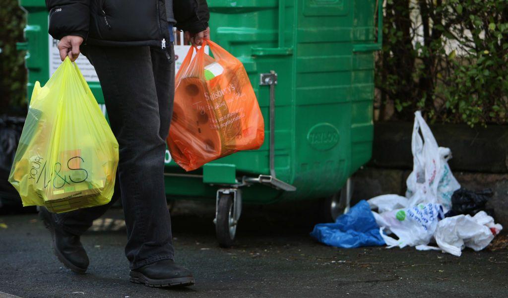 A person carrying two plastic carrier bags - one green Marks and Spencer and one orange Sainsbury's - full of groceries. In the background stands a large green bin and a pile of littered plastic bags.