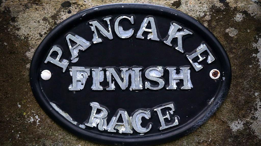 A black plaque which is weathered. In silver writing it says PANCAKE RACE FINISH.