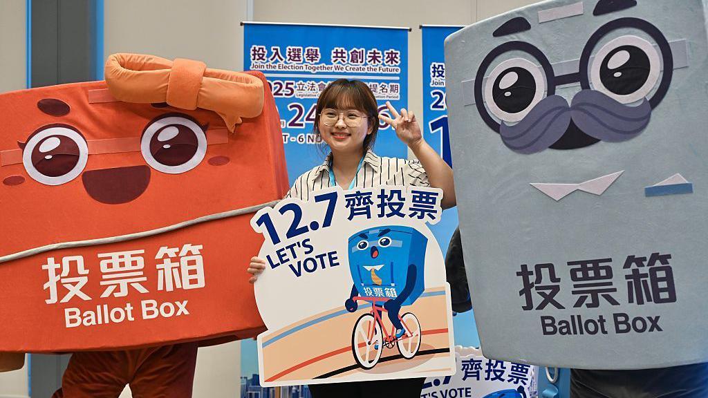 A woman in a striped shirt poses with a sign that says "12.7 Let's vote" with two life-size cartoon mascots of ballot boxes