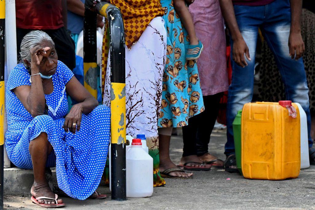 A woman in a blue spotted dress sits in a queue of people next to gasoline containers