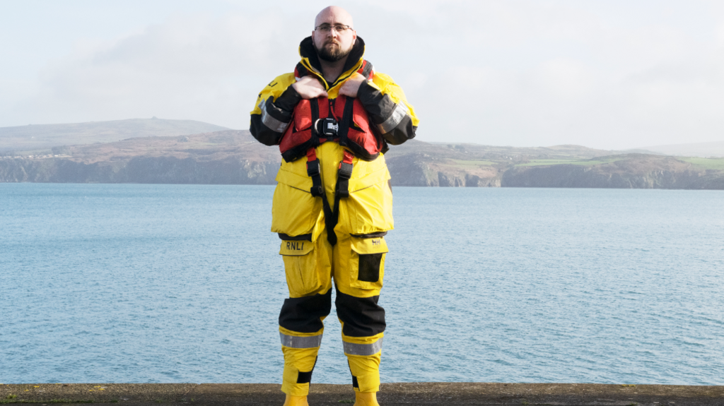 Cedwyn Rogers stands in front of the water in a yellow RNLI uniform. 