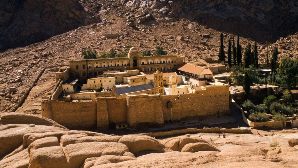 The St Catherine's Monastery as seen from the outside. It is a walled structure with several buildings inside, with the monastery's gardens attached to one side, which are green with some trees inside. The surrounding area is rocky with the ground sloping up behind the monastery at the foot of a mountain