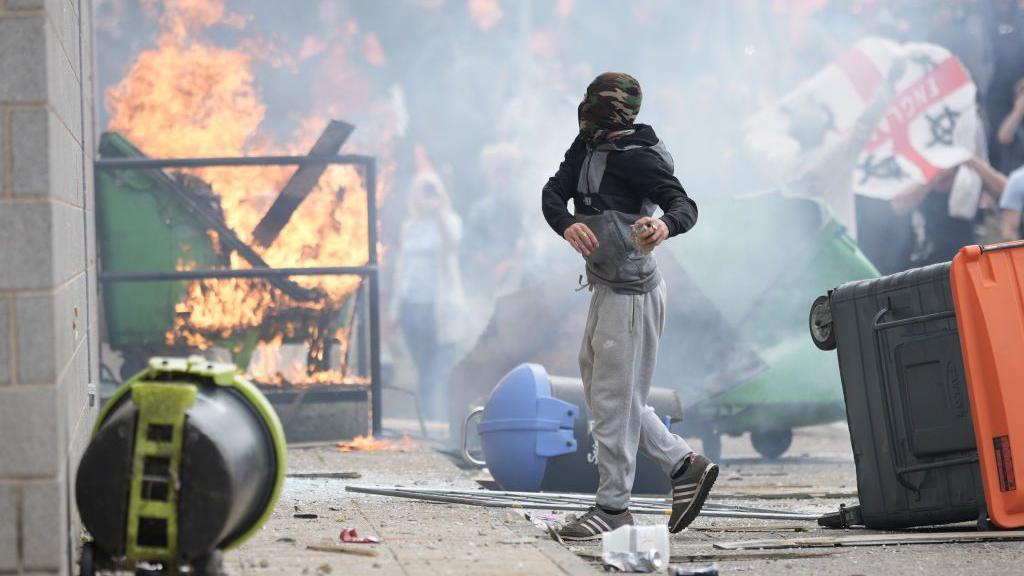 Young man in a camouflage balaclava outside a hotel in Manvers. A bin is on fire and crowds wave England flags and throw objects.