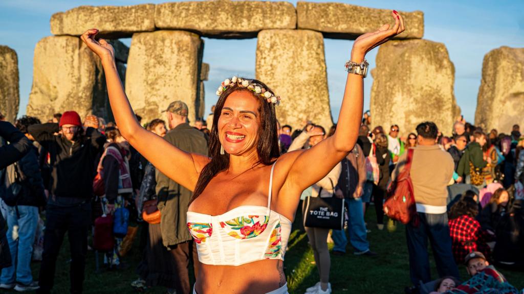 A woman stand with arms in the air, while smiling, in front of Stonehenge