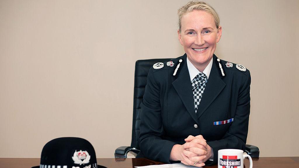 A smiling Serena Kennedy, in her police uniform, sits at a brown desk with her police hat and a Yorkshire Tea branded mug placed in front of her