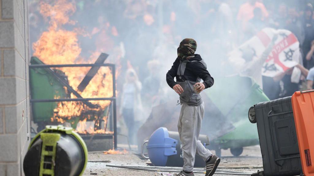 A masked man holds something in his hands as a building is set on fire