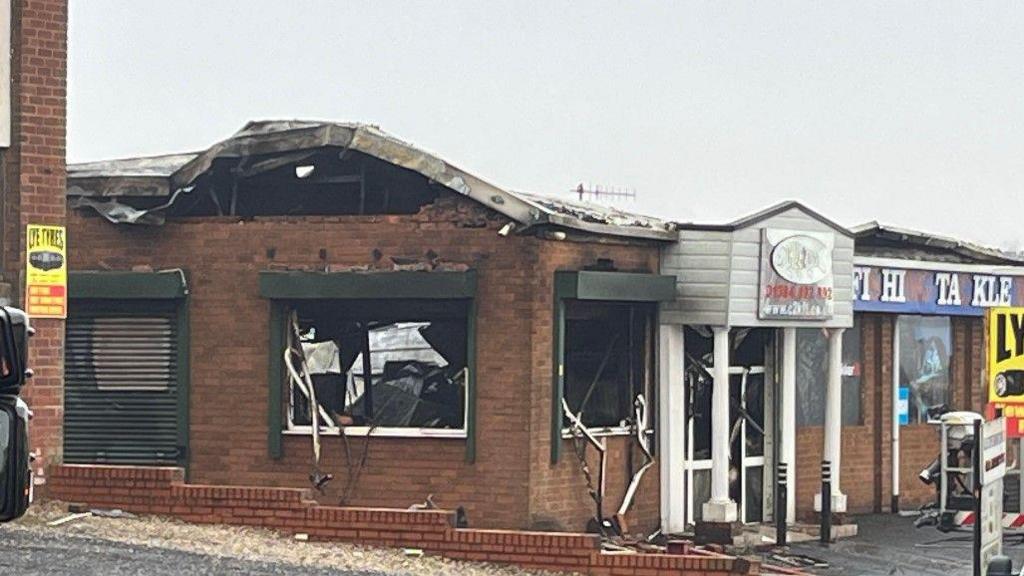 A red brick commercial building pictured from across the street in the daytime. The building is blackened in places from fire damage and there is no glass in the windows.
