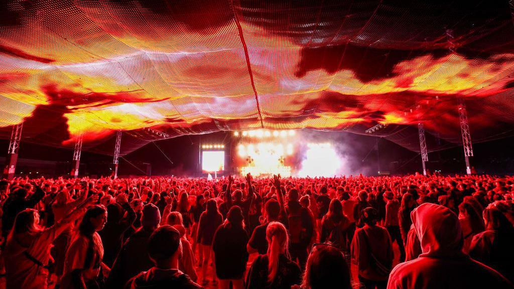 A crowd of people all facing a stage and lit up in red light. There's a light display happening on a mesh ceiling above their heads.
