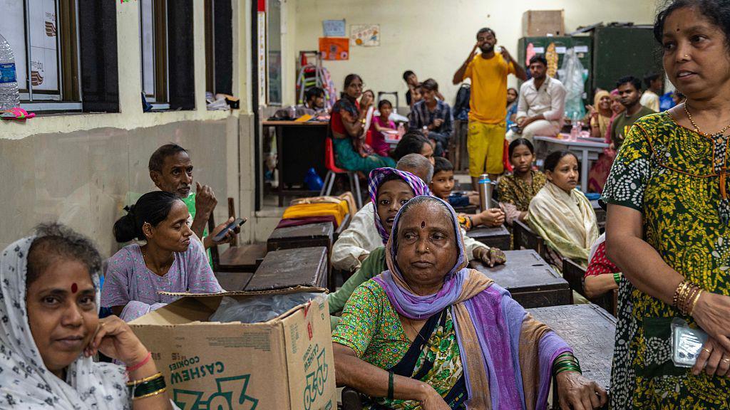 Women sitting on benches inside a classroom, which has been converted to a temporary shelter. A cardboard box is lying next to one of the women, who is wearing a green saree, and is in the centre of the picture. 