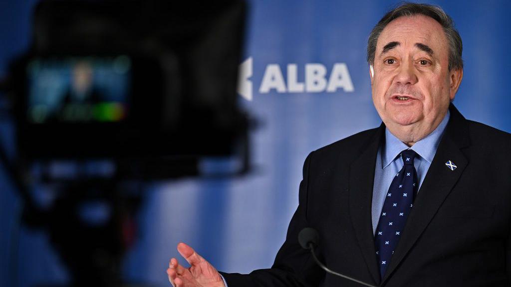 Alex Salmond standing behind a TV camera with the Alba logo visible in the background