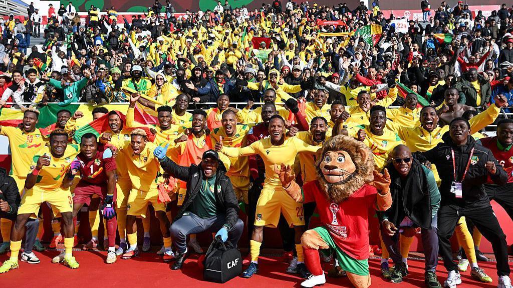 Benin footballers celebrate as a large group after winning the Afcon 2025 football match between Benin and Botswana, wearing a yellow, red and green kit alongside coaching staff in black tracksuits with a lion tournament mascot in Morocco colours and fans, many wearing yellow, with flags behind them