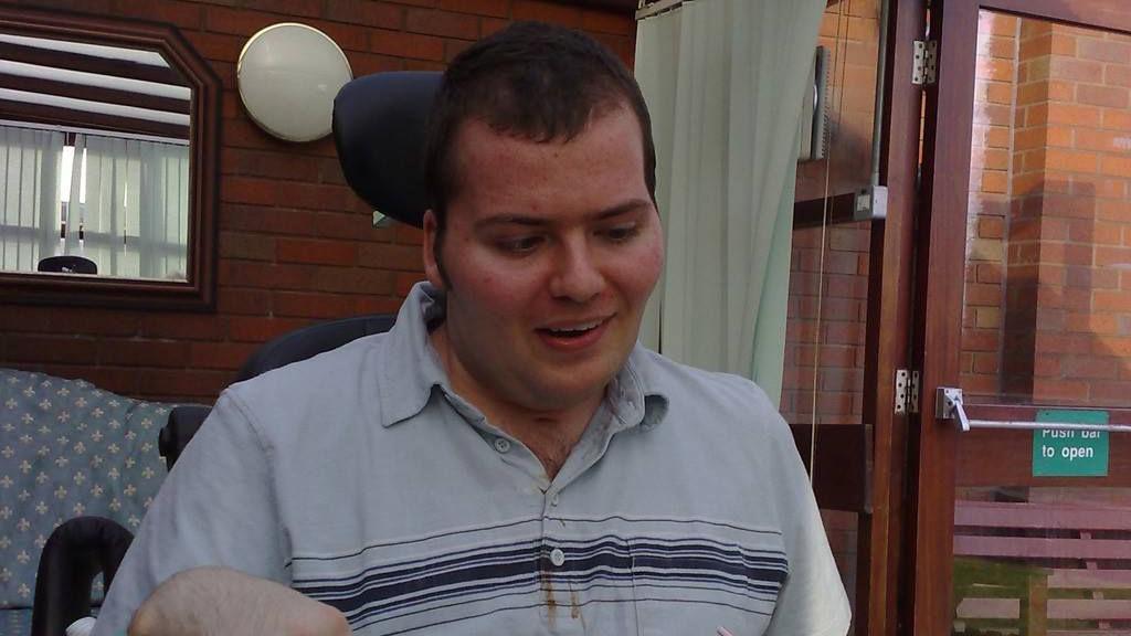 A man in a wheelchair is sitting a table. He is looking down and is wearing a grey polo shirt with a black stripes across his chest. He has short dark hair and is sitting in a conservatory built from dark red brick. 