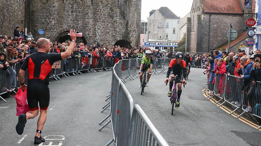 A runner passes cyclists riding in the opposite direction in the centre of Tenby as crowds of people look on from either side of the road