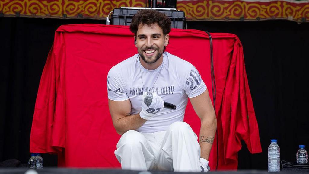 Saint Levant is wearing all white a t-shirt and trousers, he is sat down on the stage holding a mic. He has dark curly hair and brown eyes, behind his is a red tablecloth. 