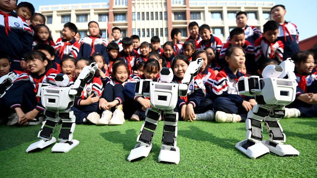 Dozens of Chinese primary school students, dressed in school uniforms, sit on a playground watching three robots dance on the ground