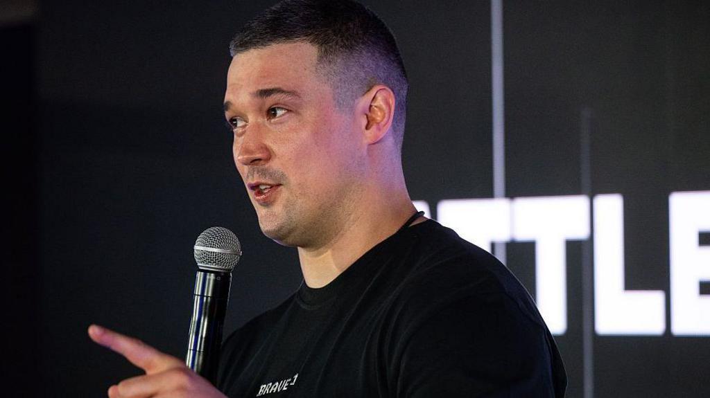 Fedorov speaking into a microphone and pointing, wearing all black