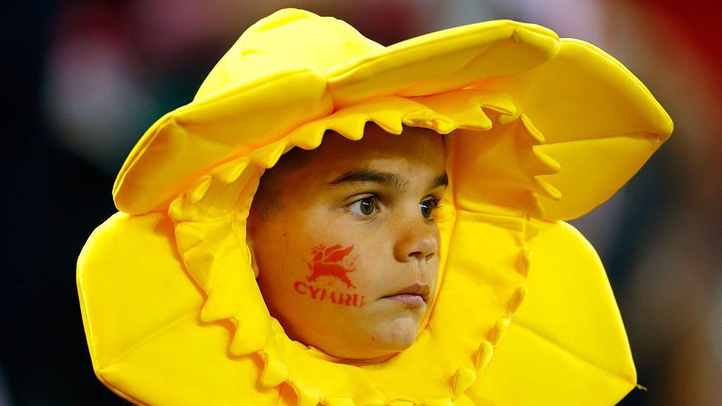 A child wears a daffodil head-dress and has a "Cymru" face-paint stamp on their cheek