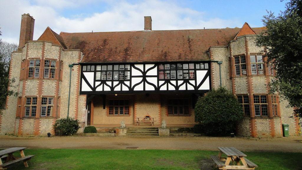 A Tudor-style mansion of flint and brick with black and white facias surrounding first floor windows in the centre of the building. Benches and grass are in front of the entrance. 